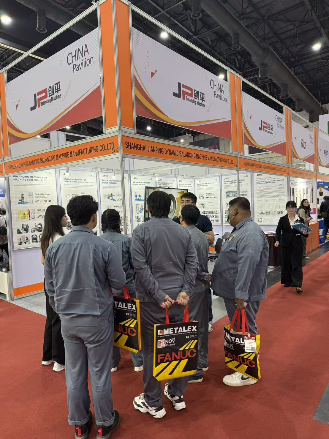 ผู้เข้าชมสวมชุดช่างถือถุง METALEX ยืนฟังทีมงานที่หน้าบูธ BY19 China Pavilion ของ JP Balancing Machine งาน METALEX 2025