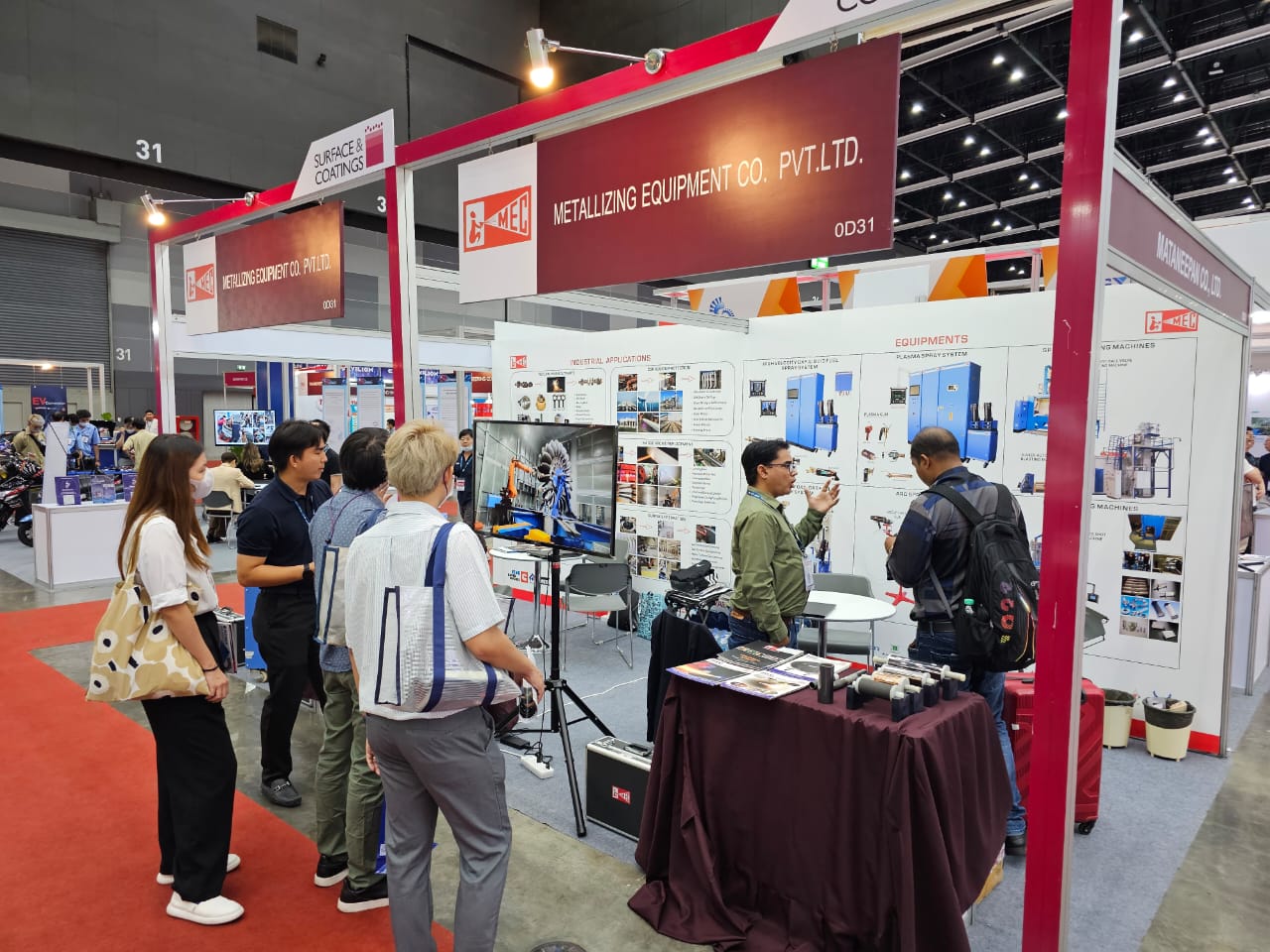 มุมมองบูธ MEC 0D31 ในงาน Surface & Coatings 2023 มีผู้เข้าชมยืนดูจอวิดีโอและโต๊ะแสดงชิ้นงาน ป้ายข้างบูธเป็นชื่อบริษัท มัทนีภัณฑ์